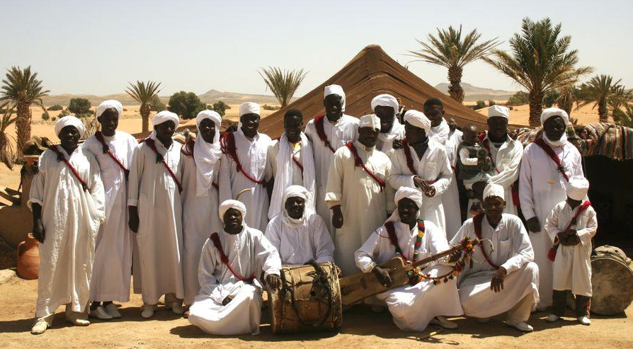 A group of men in white robes and turbans posing in the desert, some holding traditional drums and instruments