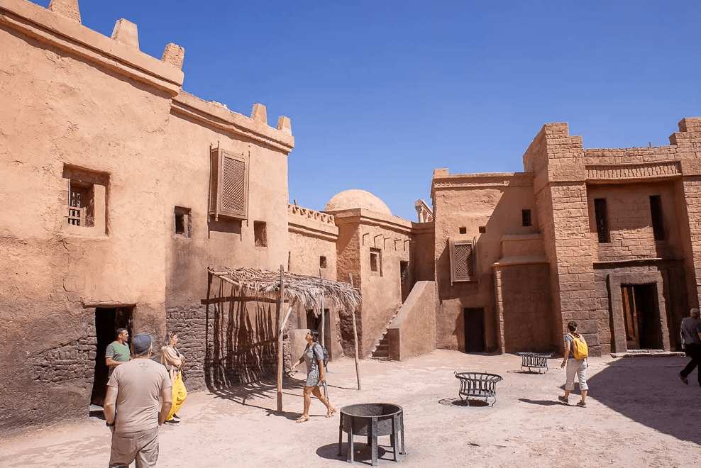 People walking through a sunlit adobe-style courtyard with tan buildings and a clear blue sky