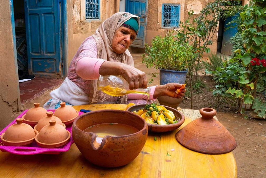 An elderly woman pours oil over a dish of vegetables in a courtyard kitchen