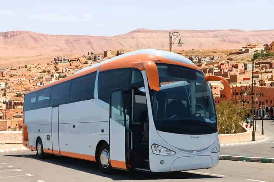 A white and orange coach bus parked on a road with desert hills and buildings in the background