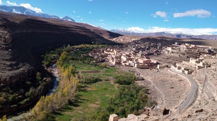 Mountain village beside a river valley with green fields and snow-capped peaks in the background