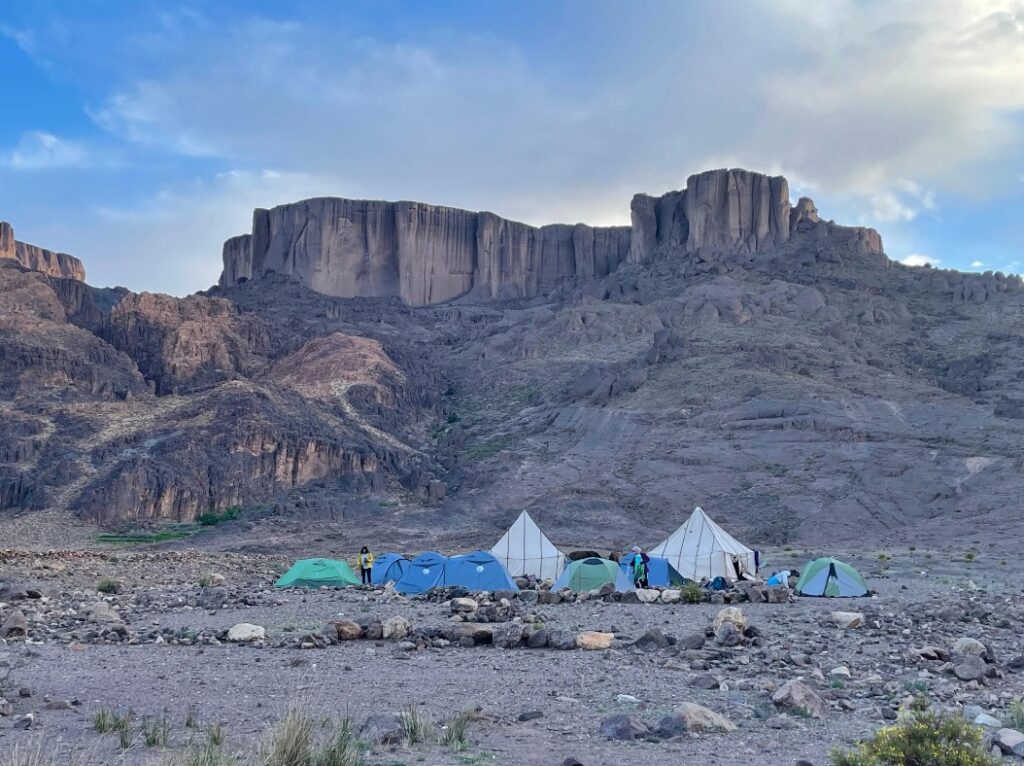 Campsite with tents in a rocky desert below steep cliffs and a cloudy sky